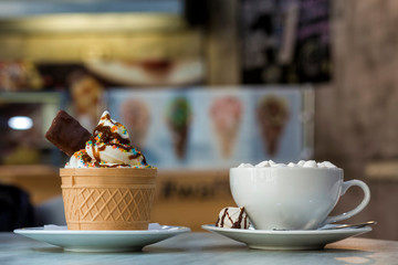 Ice cream dessert in wafer cup with chocolate cookies and and mug of coffee with marshmallows on porcelain plate on blurred colorful copy space background.