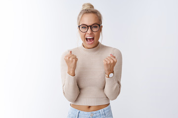 Yes we did it. Happy cheerful surprised beautiful glamour blond woman in glasses celebrating great awesome news clenching fists triumphing cheering and yelling from happiness over white wall - obrazy, fototapety, plakaty