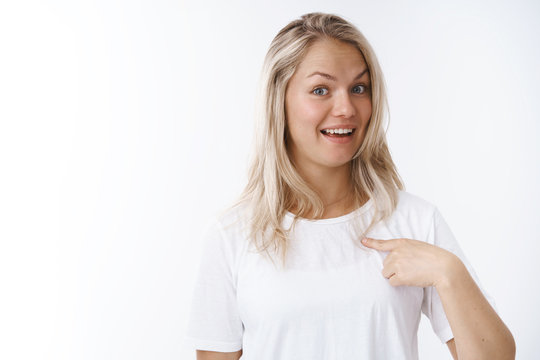 Who me. Portrait of surprised young attractive charismatic positive woman with blond hair in white t-shirt raising eyebrows in surprise smiling delighted pointing herself, being picked