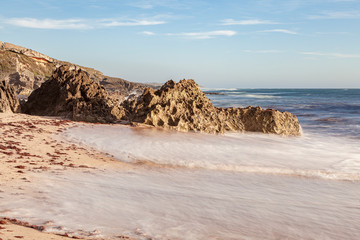 Praia com as algas vermelhas na areia trazidas pelas ondas que rebentam na areia com as rochas ao fundo do areal. Praia situada a sul de Sines em Portugal, Europa.