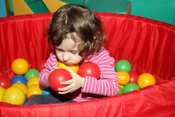 Fototapeta na wymiar Little curly cute smile girl plays in balls for a dry pool. Play room. Happiness. the child has hands full of balls
