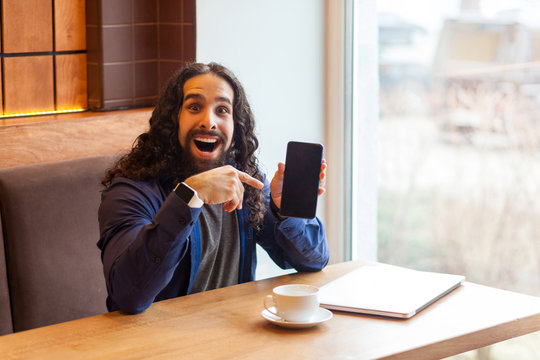 Positive amazing handsome young adult man freelancer in casual style sitting in cafe, pointing finger to empty screen phone, looking at camera and toothy smile with opened mouth. Lifestyle concept