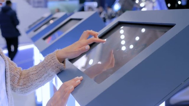 Woman using interactive touchscreen display of electronic multimedia kiosk at technology exhibition - scrolling and touching. Education, learning and technology concept