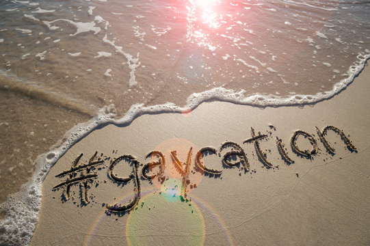 Modern gay-friendly travel message "gaycation" with a social media-friendly hashtag written in smooth sand with incoming wave on the beach