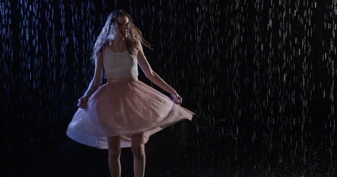 Attractive young blonde woman in white top and pastel skirt, walks into frame and dances in rain at night. Medium long shot. Slow motion 4K recorded at 60fps