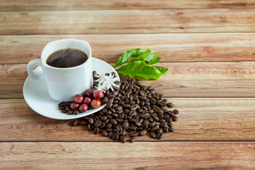 a cup of hot coffee with roasted coffee and leaves, flowers, and coffee fruit on a wooden table