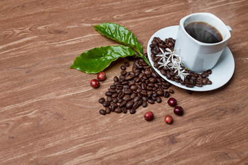 a cup of hot coffee with roasted coffee and leaves, flowers, and coffee fruit on a wooden table