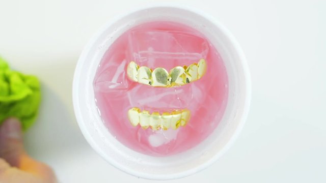 Shiny golden grillz, fake teeth jewellery sitting on colourful iced refreshing liquid. Hand with cloth cleaning surface background. Lean partydrink