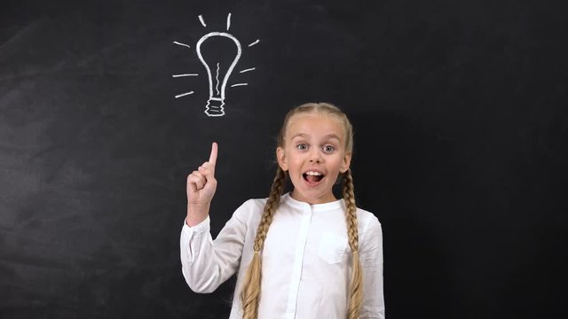 Genius schoolgirl having an idea, pointing finger at light bulb on blackboard