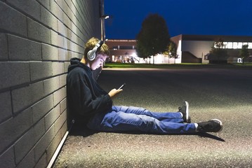 Sad teenage boy sitting on the ground against a brick wall at night. He is listening to music...