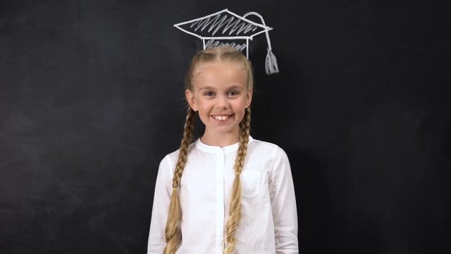 Cheerful schoolgirl smiling on camera, academic cap painted on blackboard, geek