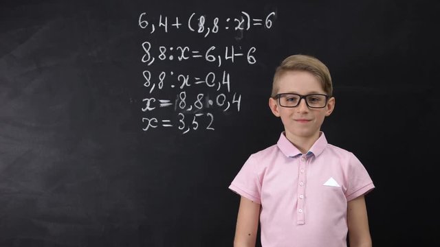 Cute nerd schoolboy in glasses standing near blackboard, math exercise written