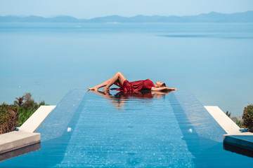 Summer. Woman model in fashion swimsuit lying on edge of infinity swimming pool with sea view. Girl...