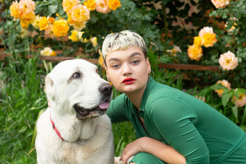 Close-up portrait of a glamorous girl with short blond hair in green dress hugging a funny alabai dog in garden against the backdrop of beautiful yellow rose bushes.