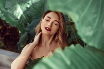 Beauty face. Woman model with bright makeup and healthy skin behind green leaf plant. Portrait of beautiful caucasian girl with big lips, closed eyes and sexy smile in tropical nature