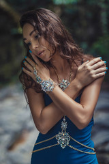 Naklejka premium beautiful long- haired brunette girl in bright blue dress and a lot of jewelry on the background of a dark forest. Bracelets, necklaces, silver