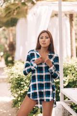 Street portrait of a young brunette asian woman  in a plaid dress standing and posing in the city park outdoor, with blurred  palm trees background. Beauty, vacation, nature, lifestyle..