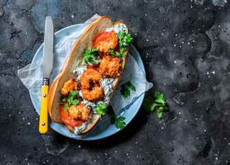 Po' boy sandwich with crispy cornmeal shrimp, tomatoes, cilantro and mayo herbs sauce on dark background, top view