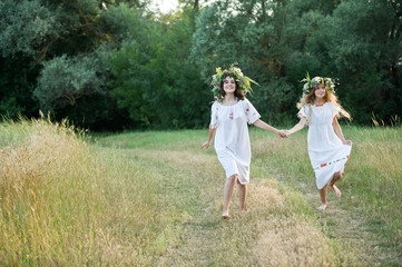 Naklejka na ściany i meble Two girls in wreaths from the field celebrate Ivan Kupala in the forest near the lake. The summer solstice