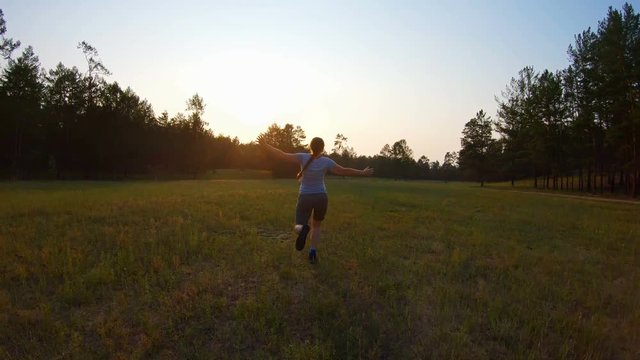 Cheerful girl, arms outstretched running across the field at sunset. Funny video, back view