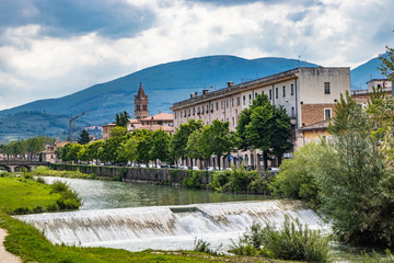Fototapeta na wymiar A view of Foligno, crossed by the river Topino, a bell tower rises above the roofs of the houses. The cloudy sky at sunset. The canal, the bridge and the trees.