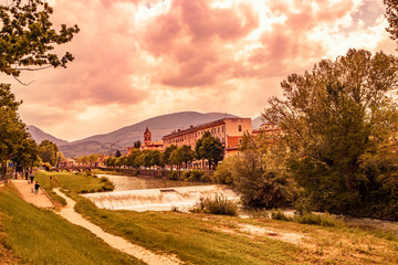 Fototapeta premium A view of Foligno, crossed by the river Topino, a bell tower rises above the roofs of the houses. The cloudy sky at sunset. The canal, the bridge, the trees. Some people walk in the park with the dog.
