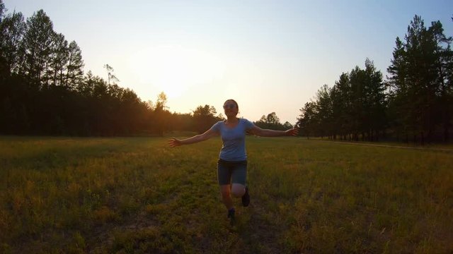 Cheerful smiling girl, arms raised running across the field at sunset. Slow motion