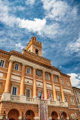 Fototapeta na wymiar The Town Hall of Foligno in Piazza della Repubblica. The beautiful facade with the colonnade, the clock, the medieval tower and the balcony with the flags of Italy and Europe. Perugia, Umbria, Italy.