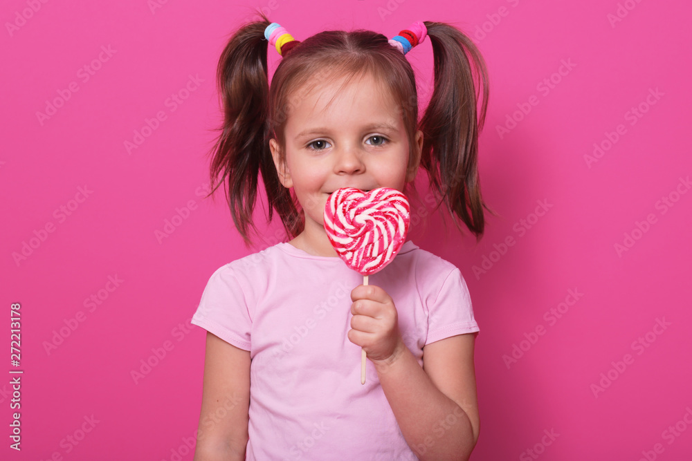 Wall mural Young pretty toddler girl kid with big sweet lollypop candy in rose casual t shirt posing isolated over pink studio background, has two funny ponitails with colourful scrinchies. Childhood concept.