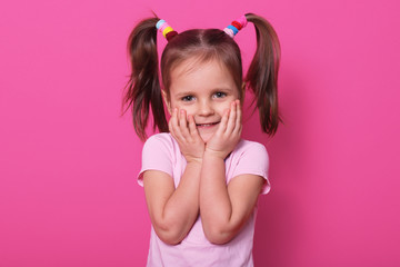 Studio shot of charming small child with two pony tails and many colourful scrunchies, expresses pleasant emotions, touches her cheeks with hands while looking directly at camera, copy spase.