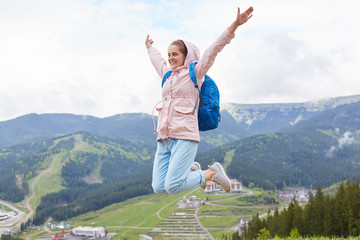 Outdoor shot of woman jumping up high, port hiking on holidays. Female wearing pink jacket and blue backpack isolated over mountains background. Traveling, adventure and active recreation concept.