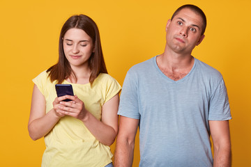 Obraz premium Studio shot of young couple, attractive female texting message on smartphone, male with thoughtful facial expression looking up, woman and man wearing casually, posing isolated over yellow studio wall