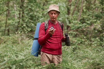 Portrait of senior man with binoculars and backpack standing in middle of forest, holding compass in his hand, looking attentively, practising to orientate in unknown place, looking concentrated.