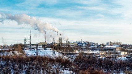 Outskirts Of Perm. In the center of the picture we see the supports of high-voltage power lines, huge Smoking pipes, plant buildings. In the foreground trees and bushes, on the ground is snow.