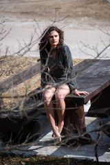 Brunette wavy hair girl in the mountains, sitting sexy on a massive wooden table. The sexy girl is hidden by a tree branch, she is wearing a short anthracite dress and has bare feet.