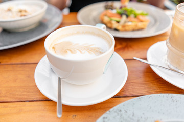 Focus in foreground on coffee cappuccino. A young woman is having Breakfast in a summer cafe.