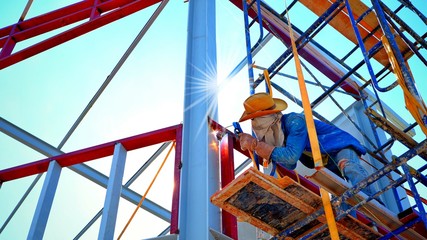 Low angle view of construction worker on scaffolding is welding metal of building structure with flare light and blue sky background, occupation and construction concept