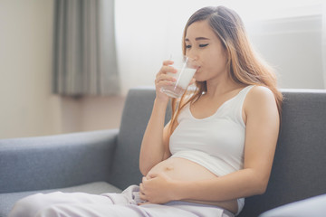 Young pregnant Asian woman is sitting on the sofa and drinking milk which is felling happy at home. To be new Mother and Father, pregnant woman concept.