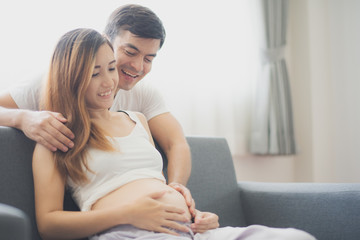 Fototapeta premium Young pregnant Asian woman is sitting on sofa with her husband are looking at her belly near window which felling happy at family's home. To be new Mother and Father, pregnant woman concept.