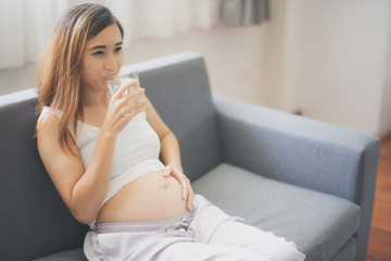 Young pregnant Asian woman is sitting on the sofa and drinking water which is felling happy at home. To be new Mother and Father, pregnant woman concept.