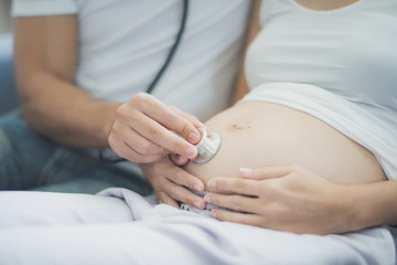 Closed up, Young pregnant Asian woman and her husband are sitting on sofa and using stethoscope to listen her belly at family home. To be new Mother and Father, pregnant woman concept.