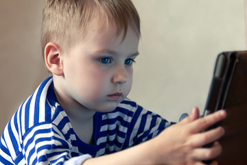 Little caucasian baby boy uses a tablet, seeing to the screen. Children time spending, computerization of youngsters. Blond hair, casual wear, indoors, close up, copy space.
