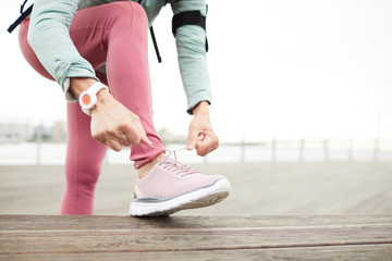 Active mature sportswoman with her leg over wooden bench tying shoelace on right sneaker before outdoor training