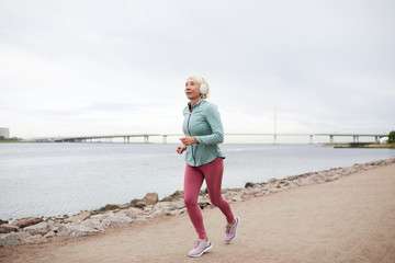 Healthy blonde contemporary sportswoman jogging along riverside while listening to music in...