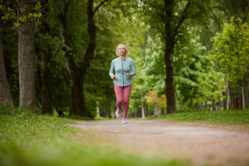 Contemporary blonde sportswoman running down path in park or natural environment among green trees on summer morning