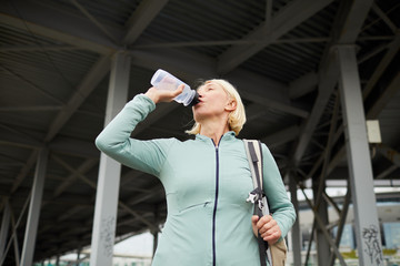 Fototapeta na wymiar Blonde mature thirsty sportswoman drinking water from plastic bottle after hard training in urban environment on summer day