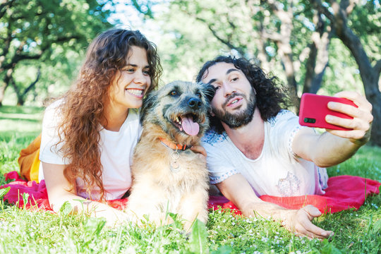 young stylish couple playing with dog in park, making selfie photo