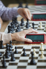 Fototapeta premium Wrinkled hand of aged experienced grandmaster in beige sweater gently pressing chess clock