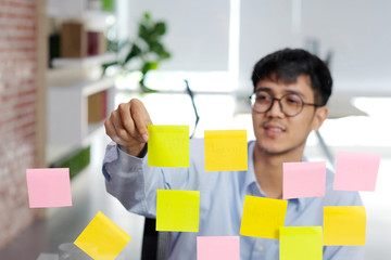 Young asian man reading sticky note on glass wall at office, business brainstorming creative ideas, office lifestyle, success in business concept