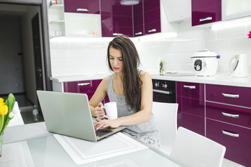 Young attractive woman working at the laptop on the kitchen. Female typing on pc indoors in the morning.